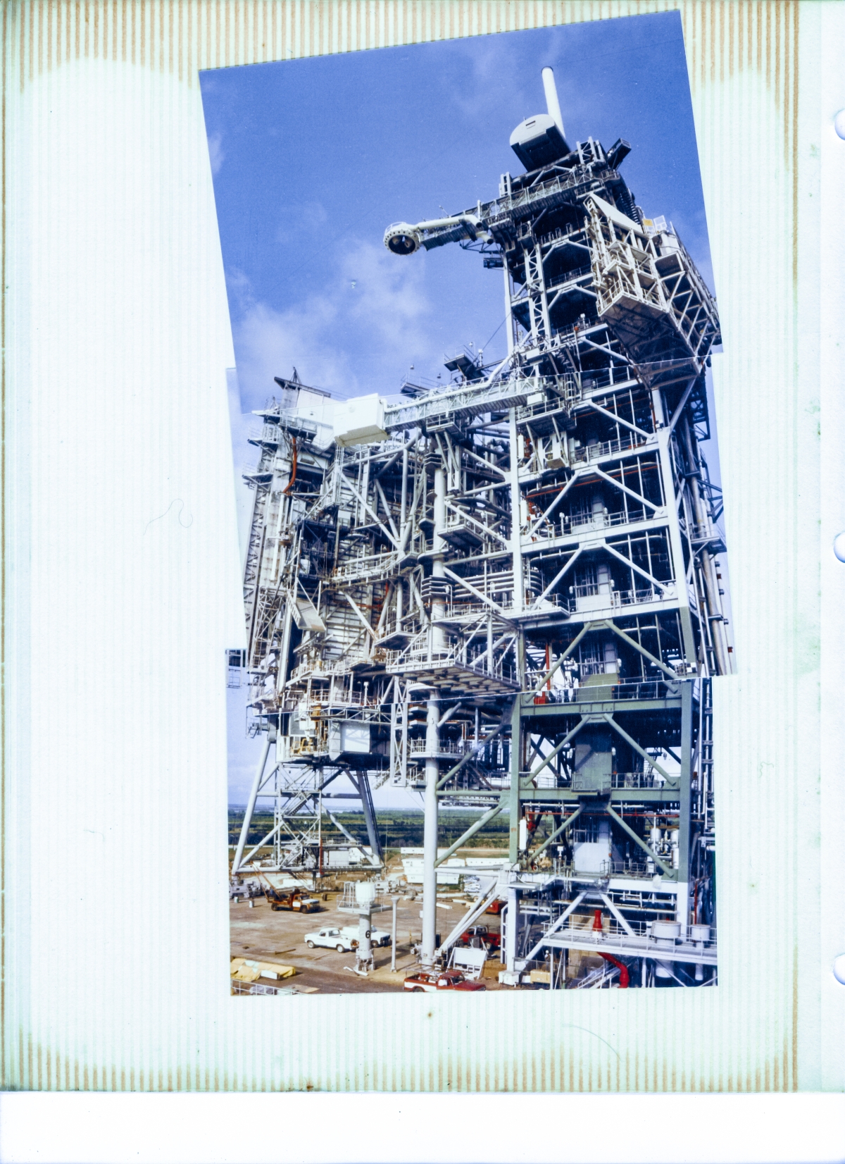 Viewed from above the pad deck at Space Shuttle Launch Complex 39-B, standing on the east MLP Access Stair Tower, this panorama of photographic prints affords an excellent overview look of the entirety of the steel structures which were required to service the orbiter prior to launch. To the left, the Rotating Service Structure hangs suspended, spanning the open spaces of the pad deck between Column Line 7 on the far side, and the Hinge Column on the near side, and the Hinge Column is tied firmly to the Fixed Service Structure, on the right side. There is much to see in this image. Much to consider.